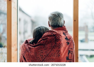 shot from behind of embraced elderly couple wrapped in a blanket looking out of a large glass wall - Powered by Shutterstock