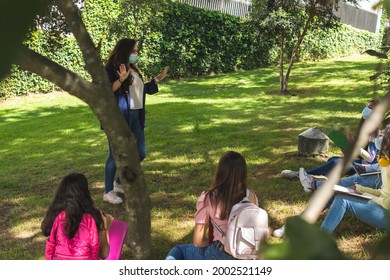 Shot From Behind A Tree Of A Latina Teacher Teaching Masked Classes Outdoors In A Park.