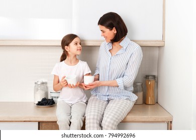 Shot of cheerful mother and daughter sit together at kitchen table, drink hot tea in morning, have pleasant friendly talk between each other. Curious girl asks something in mum during coffee break. - Powered by Shutterstock