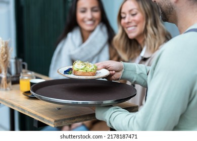 Shot Of Close Up Of Waiter Serving Toast For Brunch To Two Modern Girls Sitting On The Terrace On The Healthy Coffee Shop Terrace.