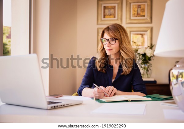 Shot of confident\
woman sitting behind her laptop and having video call while working\
from home. Home office.