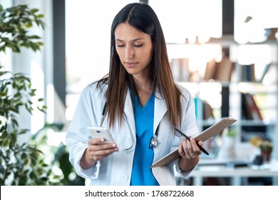 Shot Of Confident Young Female Doctor Using Her Mobile Phone While Holding A Clipboard And Standing In The Consultation.