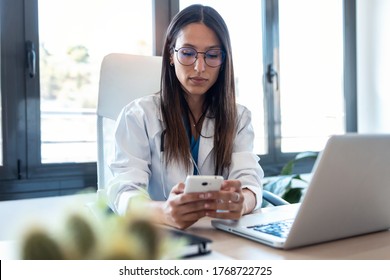 Shot of confident young female doctor using her mobile phone while working on laptop in the consultation. - Powered by Shutterstock