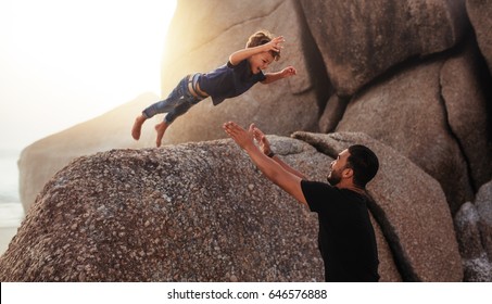 Shot of cute little boy leaping into his fathers arms from a big rock. Father and son enjoying summer holidays at the beach.