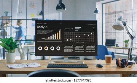 Shot of a Desktop Computer in the Creative Modern Office. Monitor Screen Shows Company Growth Data with Graphs, Charts, Software UI. In the Background Young Professional Looking for a Book.