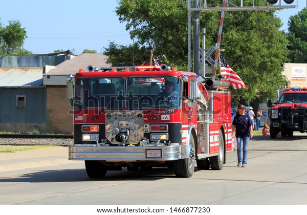 A shot of an Ellsworth Co\
Fire Truck in a Parade in Wilson Kansas USA. With the US Flag,blue\
sky,green tree\'s, and people in the parade. That\'s on\
7-27-2019.