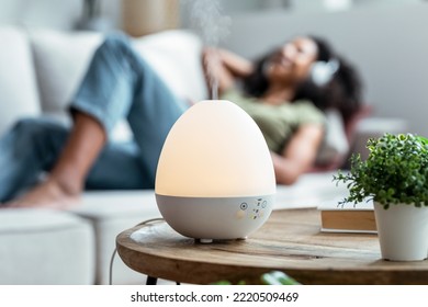 Shot of essential oil aroma diffuser humidifier diffusing water articles in the air while woman listening music lying on coach. - Shutterstock ID 2220509469
