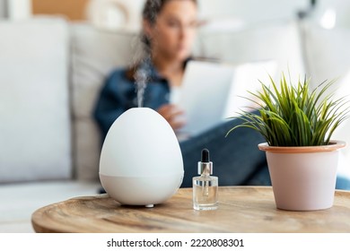 Shot of essential oil aroma diffuser humidifier diffusing water articles in the air while woman reading a book sitting on coach. - Shutterstock ID 2220808301