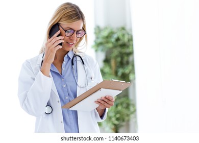 Shot Of Female Doctor Using Her Mobile Phone While Reviewing Some Papers In The Consultation.