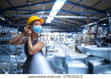 Shot of female factory worker in uniform and hardhat putting on face mask in industrial production plant. People working during COVID-19 pandemic.