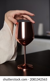 Shot of a female hand with a bronze steel wine glass.The fingers are on the top of the glass. The goblet stands on a black table. There is a dark gray wall and different items at the background. - Shutterstock ID 2081342563