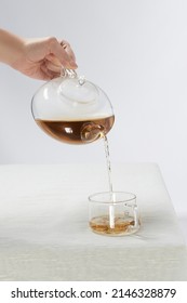 Shot of a female hand pouring tea from a teapot into a transparent cup with an original handle. The tea set is located on the white table on the white background.