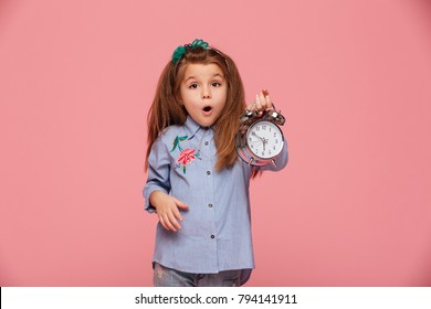 Shot Of Female Kid Posing On Camera With Eyes And Mouth Wide Open, Holding Clock Nearly 6 Being Shocked Or Shaken Up Over Pink Background