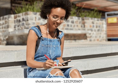 Shot of female student listens audio book with earphones and mobile phone, writes some records and details in diary, poses at stairs outdoor, prepares for seminar, uses internet and technology.