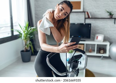 Shot Of Fitness Woman Using Mobile Phone While Training On Exercise Bike At Home.