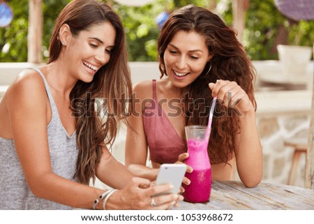 Shot of glad female models choose photo to update in social networks, view images on smart phone with happy expressions, drink fresh smoothie in terrace bar. Women tourist book hostel online