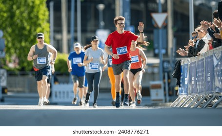 Shot of a Group of Diverse People Running in a Marathon and Waving to their Loved Ones and Supporters in the Audience. Runners Participating in a Charity Run to Raise Money a Cause - Powered by Shutterstock