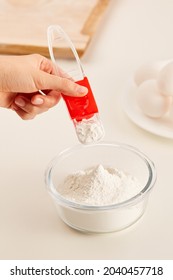 Shot of hand that holds a red measuring spoon with a movable scale above a glass bowl with flour. Bowl stands on a white table. Wooden cutting board and a white plate with eggs on the background.