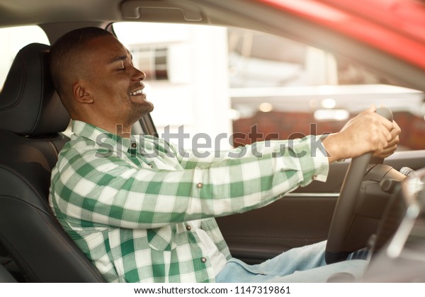 Shot of a handsome African man smiling with his\
eyes closed enjoying sitting in a new comfortable automobile at the\
dealership. Male customer choosing car to buy. Lifestyle, driving,\
safety concept