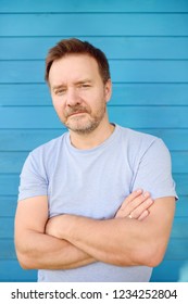 Shot Of Handsome Mature Man Crossing Hands And Look At Camera. Male Portrait With Blue Wooden Wall On Background.