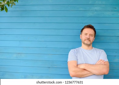 Shot Of Handsome Mature Man Crossing Hands And Look At Camera. Male Portrait With Blue Wooden Wall On Background.