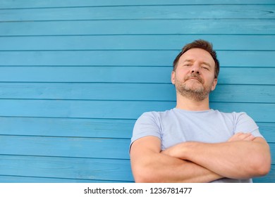 Shot Of Handsome Mature Man Crossing Hands And Look At Camera. Male Portrait With Blue Wooden Wall On Background.