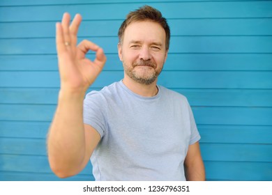 Shot Of Handsome Mature Man Looking At Camera And Showing OK Gesture. Male Portrait With Blue Wooden Wall On Background. Concept Of Success.