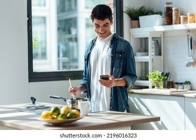 Shot Of Handsome Young Man Cooking And Using His Mobile Phone In The Kitchen At Home.