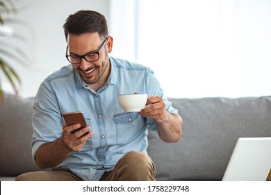 Shot of a handsome young man drinking coffee at home. Happy man drinking a cup of coffee at home. Shot of a young man using a smartphone and having coffee on the sofa at home - Powered by Shutterstock