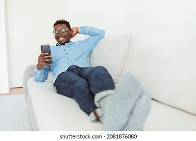 Shot of a handsome young man using his cellphone while relaxing on the couch at home. Young black american guy, browsing on his smart phone, sitting on cozy beige sofa at home, in denim casual wear.
