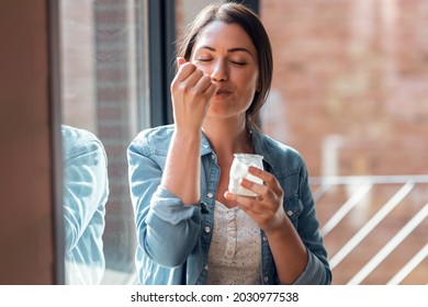 Shot of happy beautiful woman eating yogurt while standing in living room at home. - Powered by Shutterstock