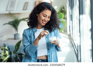 Shot of happy beautiful woman eating yogurt while standing in living room at home.