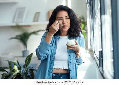 Shot of happy beautiful woman eating yogurt while standing in living room at home.