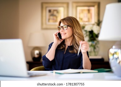 Schuss eine glückliche Geschäftsfrau, die am Schreibtisch hinter ihrem Laptop sitzt und mit jemandem auf ihrem Handy spricht, während sie von zu Hause aus arbeitet. Heimbüro.