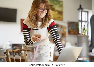 Shot Of A Happy Coffee Shop Owner Using Mobile Phone And Laptop While Standing At Desk And Manage Her Small Business.