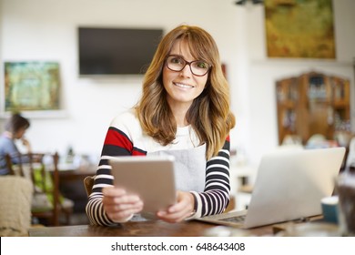 Shot Of A Happy Coffee Shop Owner Using Digital Tablet And Laptop While Sitting At Desk And Manage Her Small Business.