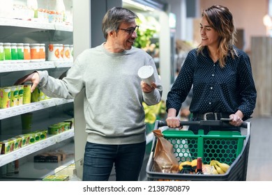 Shot Of Happy Couple Talking While Walking With Shopping Cart And Taking Products From Shelf At The Grocery