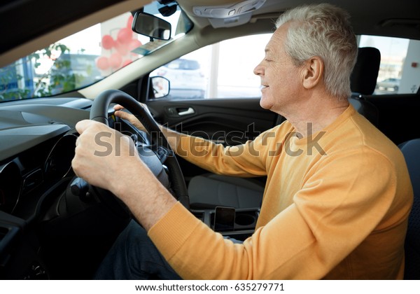 Shot of a happy elderly male customer sitting in a\
new car at the dealership examining interior of an auto touching\
dashboard smiling comfort luxury happiness vehicle transportation\
quality modern