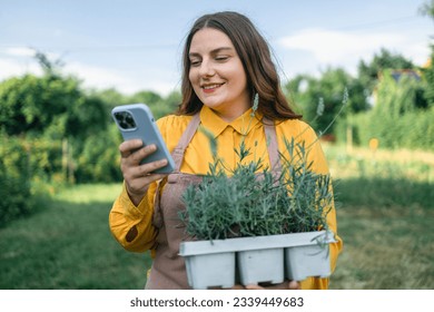 Shot of an happy female Caucasian farmer using a smartphone and holding a plastic pot of lavender flower plants to plant in the ground. Gardener using mobile phone  - Powered by Shutterstock