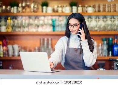 Shot Of Happy Female Coffee Shop Owner Standing In The Coffee Shop And Working. Young Waitress Using Mobile Phone And Laptop. 