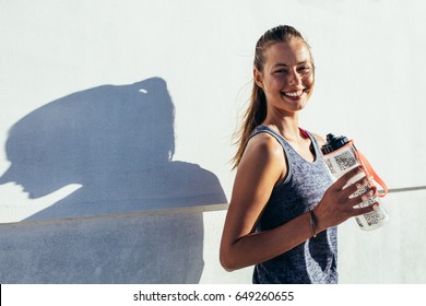 Shot of happy female runner standing outdoors holding water bottle and smiling. Fitness woman taking a break after workout.