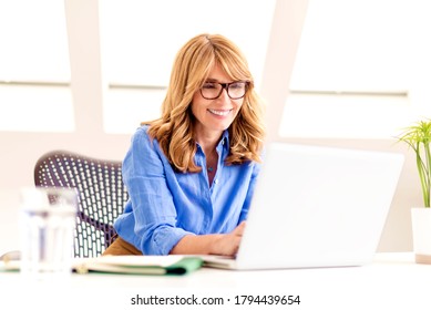 Aufnahme einer glücklichen, reifen Geschäftsfrau, die am Schreibtisch sitzt und an Laptop im Büro arbeitet.