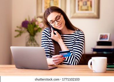 Shot of happy woman holding a bank card in her hand and making a phone call while sitting in front of laptop and shopping online. 