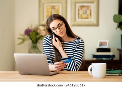 Shot of happy woman holding a bank card in her hand and making a phone call while sitting in front of laptop and shopping online.  - Powered by Shutterstock