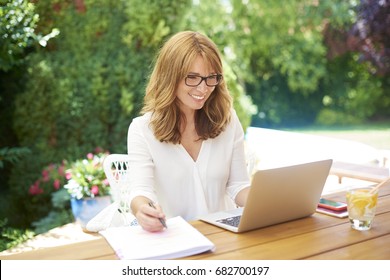 Shot of a happy woman sitting on balcony and writing something while working on laptop at home.  - Powered by Shutterstock