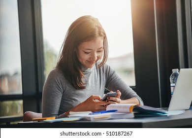 Shot Of Happy Young Female Student Sitting At Table Reading Text Message On Her Cell Phone. Asian Girl Using Mobile Phone At College Library.