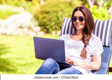 Shot Of Happy Young Woman Sitting On Sunbed In The Garden And Using Her Laptop.