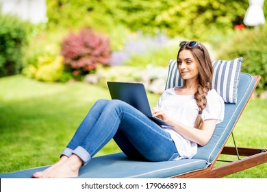Shot of happy young woman sitting on sunbed in the garden and using her laptop.