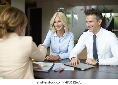 Shot Of An Investment Manager Shaking Hands With Smiling Woman While Consulting With Middle Aged Couple About Financial Savings. 