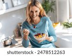 Shot of kind beautiful woman eating a healthy fruit bowl while sitting on the table in the kitchen at home